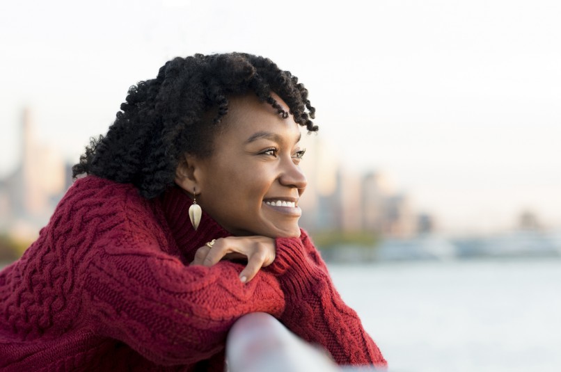 Close up portrait of a young happy african woman leaning on the banister of a bridge near river. Happy young african woman at river side thinking about the future. Smiling pensive girl looking across river at sunset.
