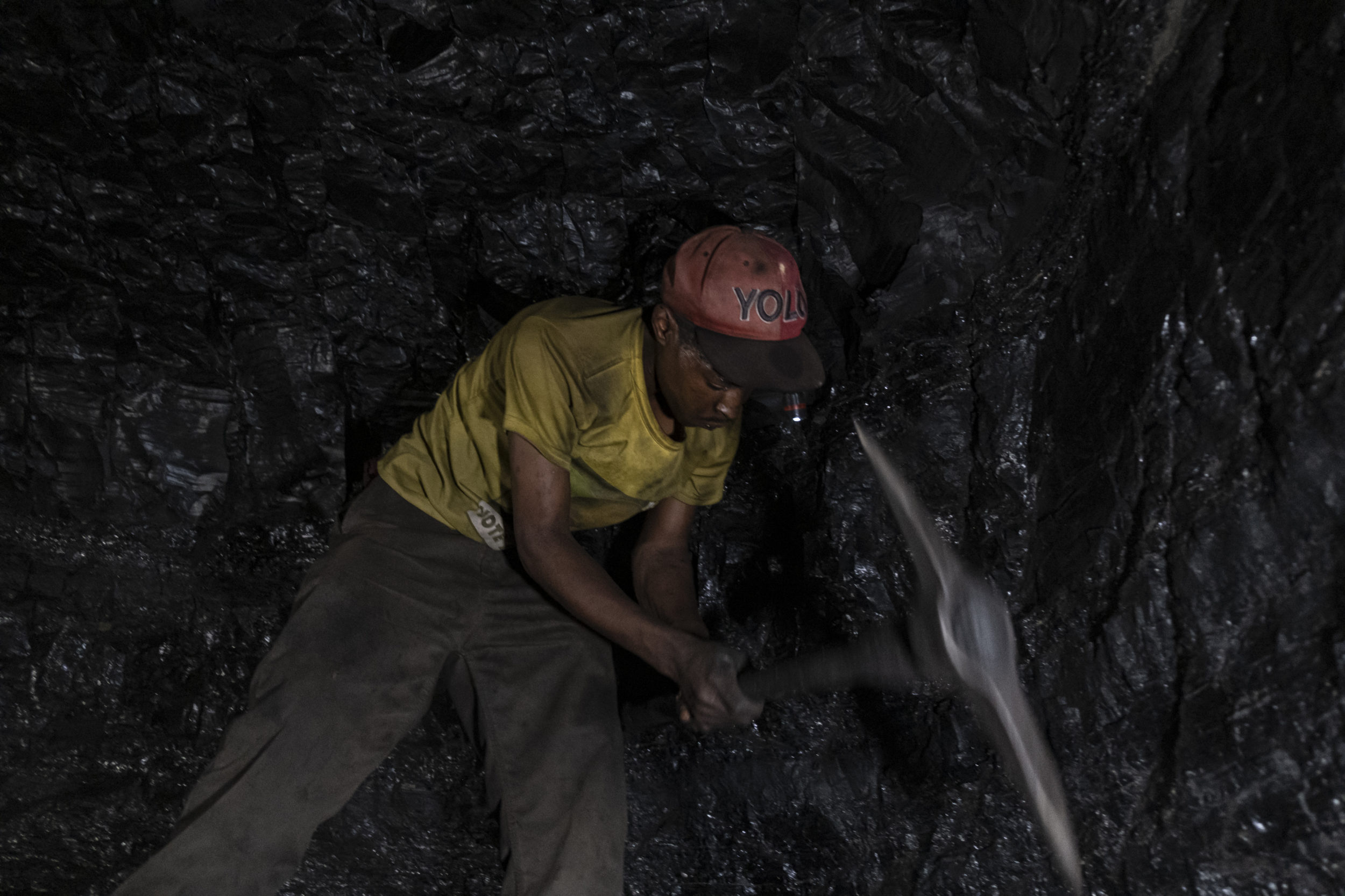 Bonginkosi Mhlanga swings his axe pick to the wall of coal at the abandoned Goldview coal mine in Ermelo, Mpumalanga on 20 April 2021. After skidding down 82 steps and making his way through a narrow tunnel, Bonginkosi reached a black vein in the rock some two meters wide where he would remain overnight breaking off and collecting coal, rarely allowing himself to withdraw from labour. Picture: Emmanuel Croset/AFP