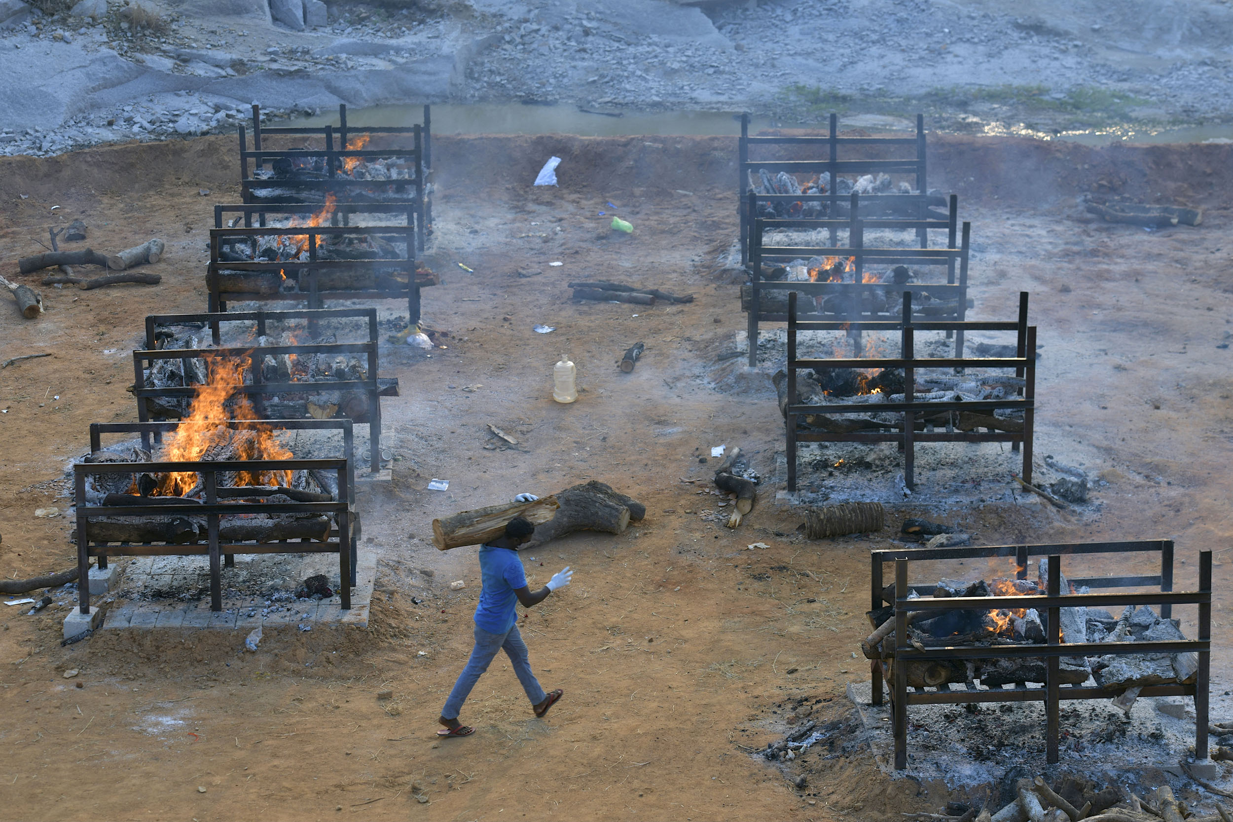 An undertaker carries a wooden log amidst burning pyres of the victims who died of Covid-19 coronavirus at an open air crematorium set up for the coronavirus victims inside a defunct granite quarry on the outskirts of Bangalore on May 1, 2021. (Photo by Manjunath Kiran / AFP)